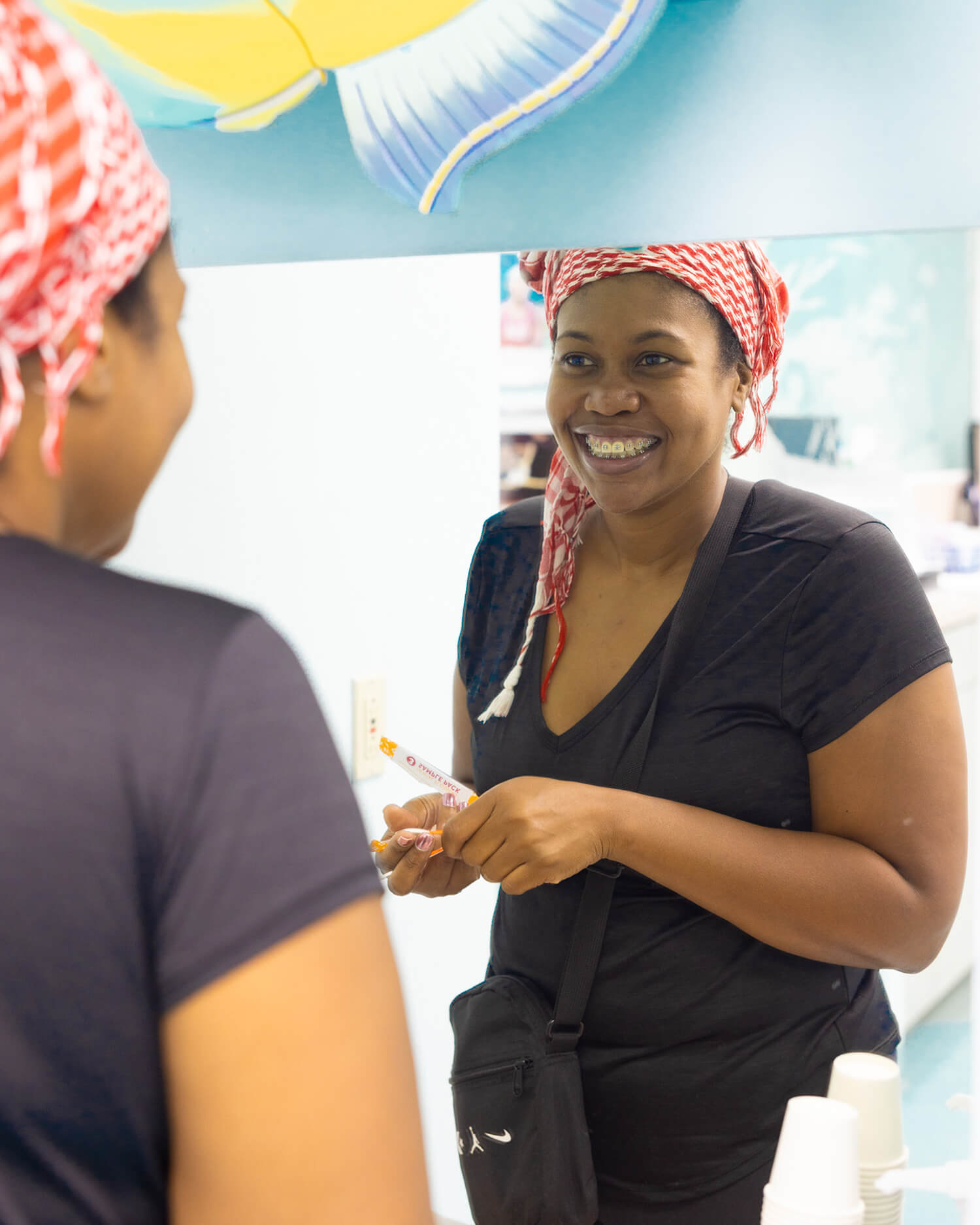 Woman in black shirt wearing pink headwrap smiles while speaking with patient in orthodontic office setting.