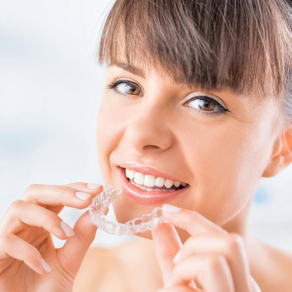 Young person smiling while holding clear orthodontic aligner against bright background.