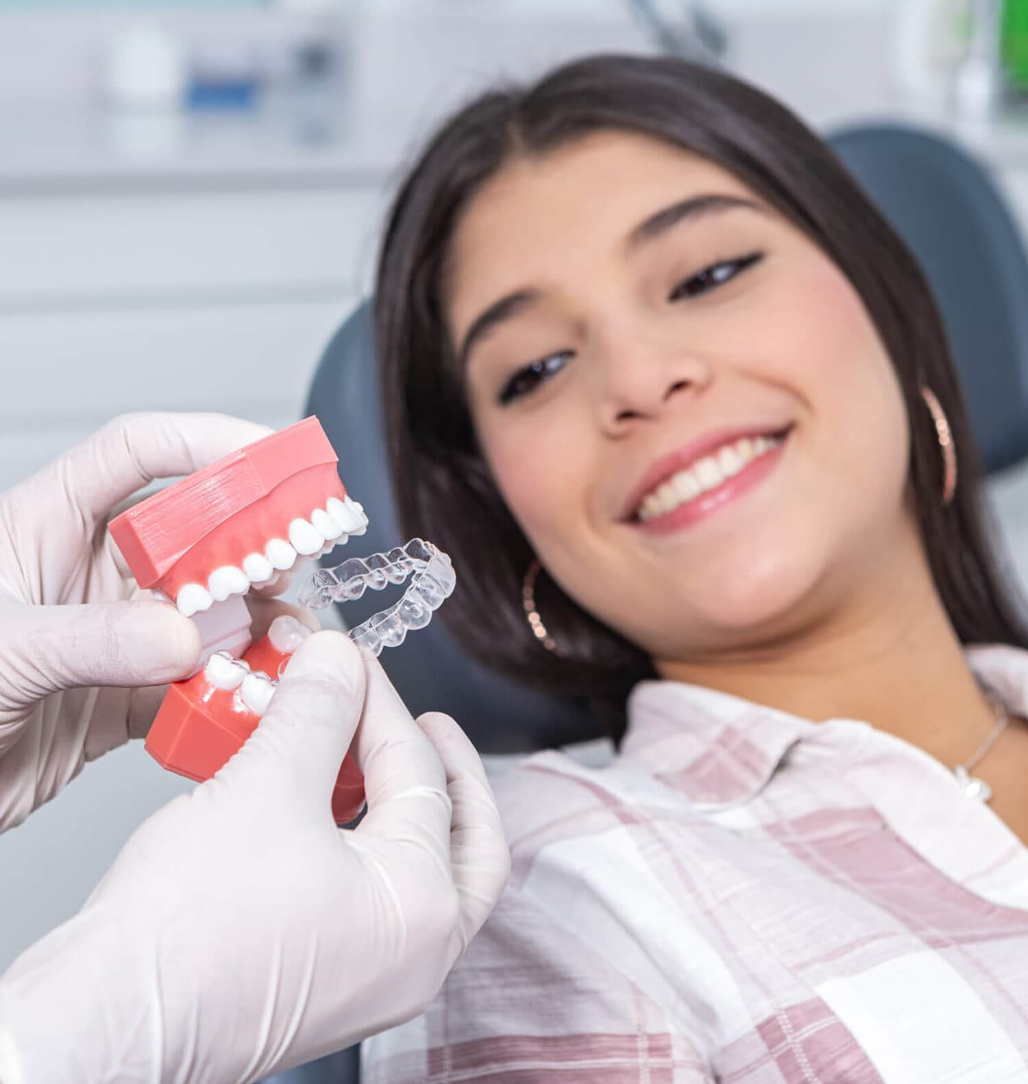 Young woman in dental office holding orthodontic model, smiling at camera with braces visible.