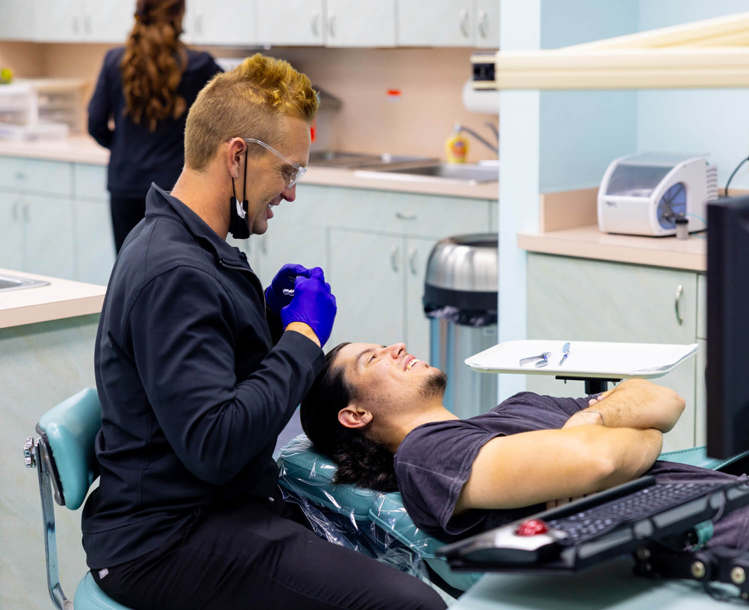 Orthodontist examining patient's teeth in modern dental office with equipment visible in background.
