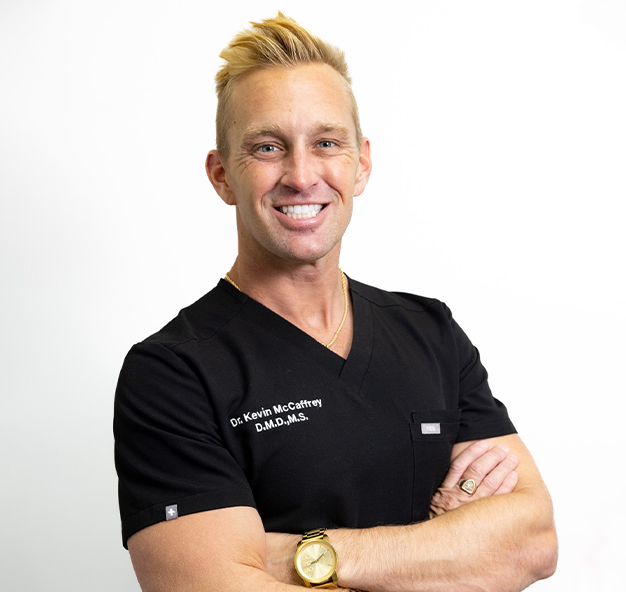 Professional orthodontist smiling with arms crossed wearing black scrubs against white background.