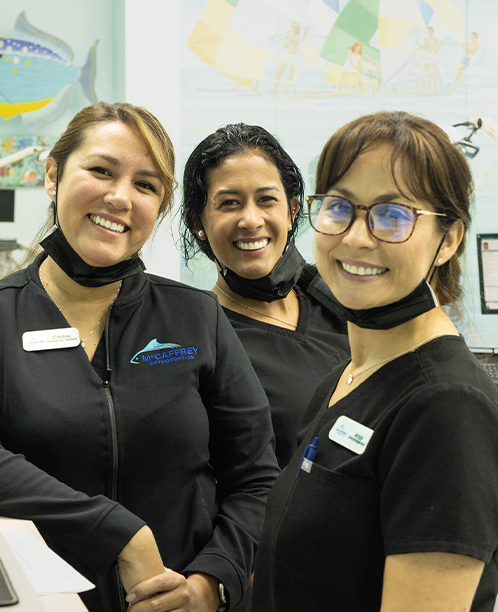 Three smiling female staff members in black uniforms pose together in an orthodontic office setting.
