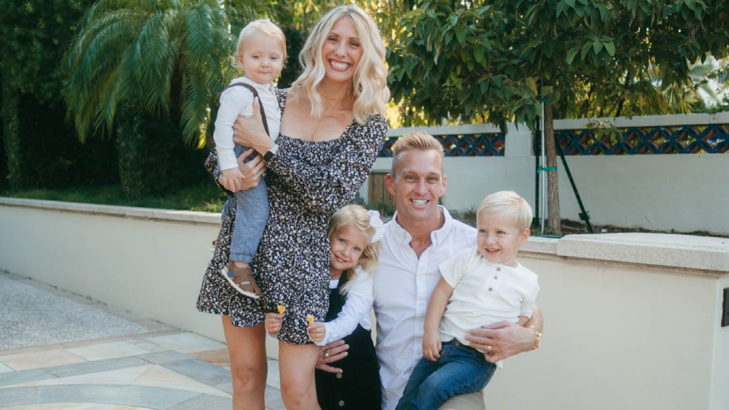 Family of five posing together outdoors on a sunny patio with white walls and trees.