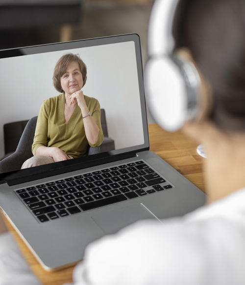 Woman on video call with patient wearing headphones at wooden desk.